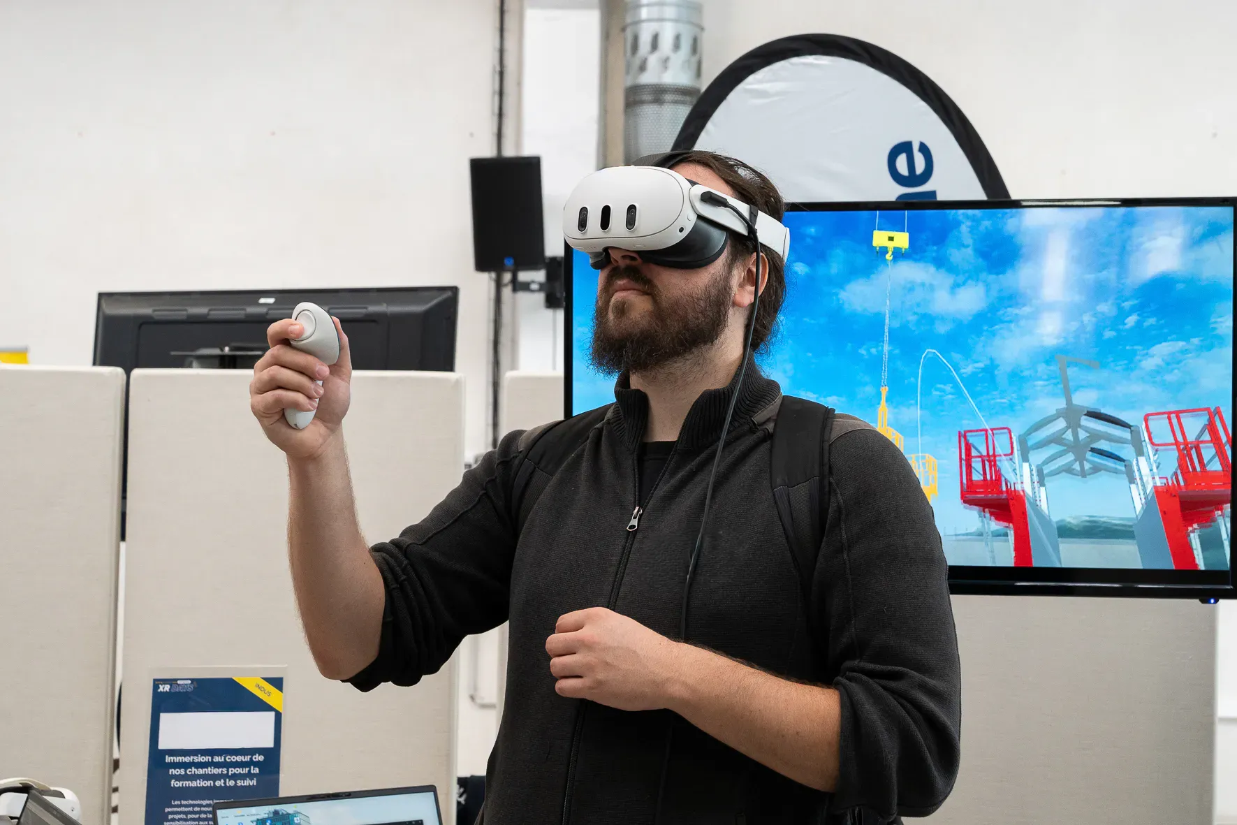 Un participant en casque de réalité virtuelle ; la même scène de chantier s’affiche sur l’écran derrière lui.